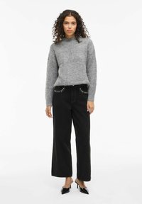 Sivi pahuljasti džemper, visoki ovratnik, duge rukave, uparen s širokim crnim trapericama sa zakovice na džepovima i crnim cipelama s potpeticama.