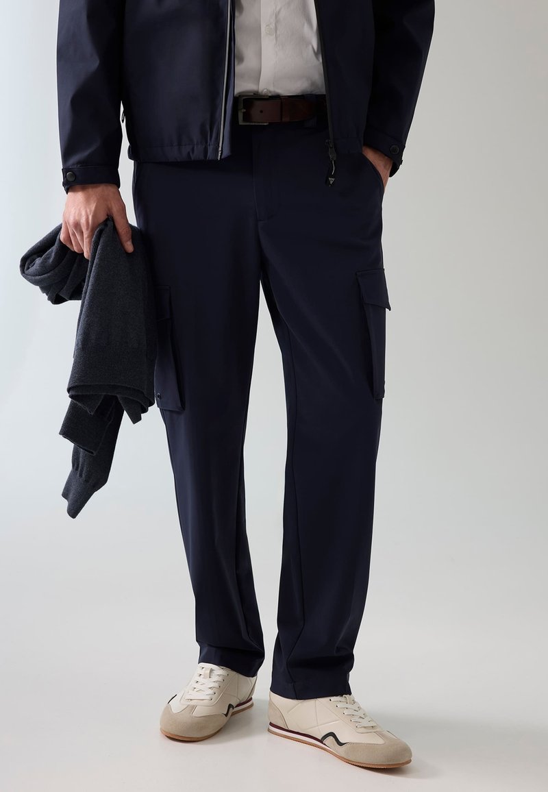 Homme portant un pantalon cargo bleu marine, des baskets blanches et une veste foncée tenant un pull foncé plié dans une main.