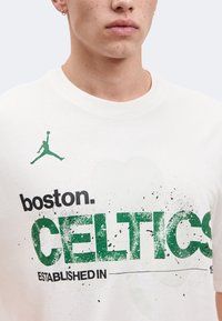 Κοντινό πλάνο ενός ατόμου που φοράει λευκό πουκάμισο με πράσινο και μαύρο κείμενο "Boston Celtics Established" και πράσινο λογότυπο Jumpman στο στήθος.