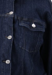 Mørkeblå denimjakke med sølvfargede metallknapper, to brystlommer og synlige sømdetaljer. Glatt tekstur og klassiske designelementer.