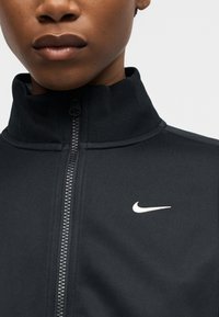 Κοντινό πλάνο ενός ατόμου που φοράει μαύρο μπουφάν με φερμουάρ και ψηλό γιακά, με λευκό λογότυπο Nike Swoosh στο στήθος.