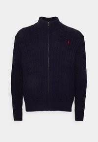 Marinblå kabelstickad zip-up kofta med hög krage, ribbade manschetter och nederkant, med en liten röd logotyp på vänster sida.