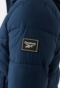 Granatowa pikowana kurtka z teksturowanej tkaniny, ze szwami oraz czarną prostokątną naszywką z logo Reebok na ramieniu.