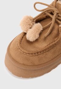 Pantof cu toc rotund din suede maro, având două pompoane de culoare bej deschis deasupra. Talpă groasă din cauciuc cu margini texturate pentru aderență.