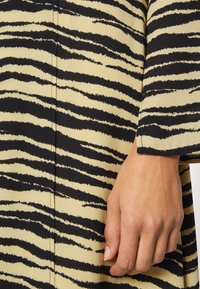 Το φόρεμα διαθέτει μοτίβο τιγρέ ρίγες σε μαύρο και μπεζ χρώμα, με ομαλή υφή· ένα χέρι είναι εμφανές κοντά στο μανίκι, αναδεικνύοντας το ύφασμα.