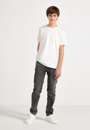 Poika seisoo valkoinen lyhythihainen t-paita, haalistuneet mustat farkut ja valkoiset tennarit päällään, yksinkertaisen vaalean taustan edessä.