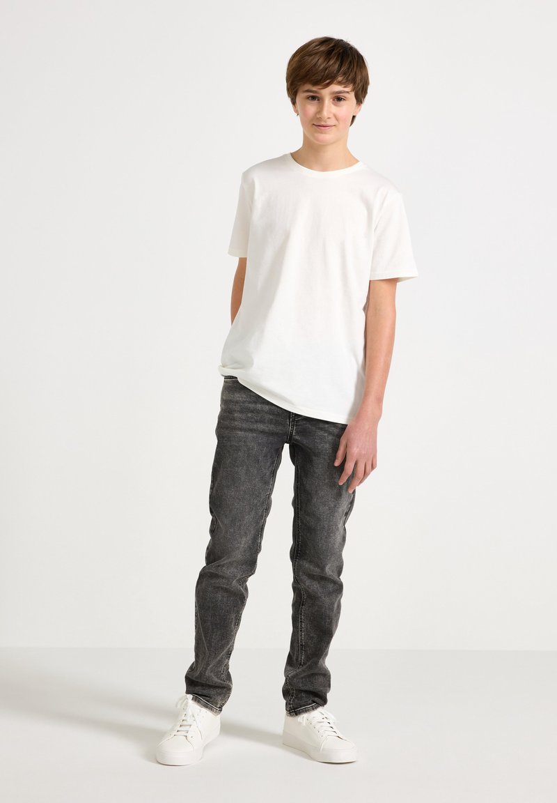 Jongen staand, draagt een wit shirt met korte mouwen, verwassen zwarte spijkerbroek en witte sneakers, tegen een effen lichte achtergrond.