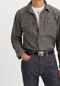 Camicia grigia con bottoni e due tasche sul petto, abbinata a jeans in denim scuro e una cintura nera con una fibbia rettangolare in argento.