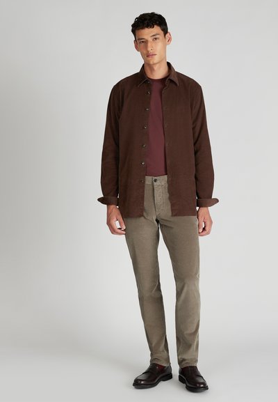 Homme portant une chemise marron à boutons sur un t-shirt bordeaux, un pantalon en velours côtelé beige et des mocassins marron foncé, se tenant devant un fond uni.