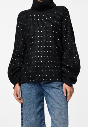 Zwarte gebreide trui met hoge nek, voorzien van zilveren studs in een ruitpatroon en puffmouwen. Gecombineerd met denim jeans.