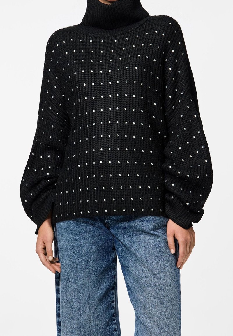 Svart stickad tröja med hög krage, prydd med silverfärgade nitar arrangerade i ett rutmönster och puffärmar. Matchad med jeans.