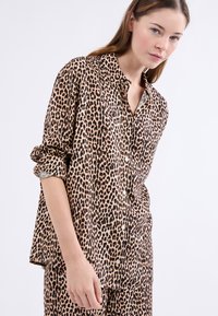 Beige ja must leopardimustriga nööpidega särk, mille varrukad on üles keeratud ja pind on sile. Esiküljel on nööbid.