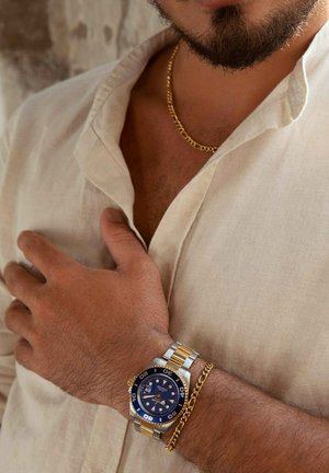 Zilveren en gouden horloge met een blauwe wijzerplaat, ronde face en glow-in-the-dark markeringen; gedragen met een gouden schakelarmband tegen een lichtgekleurde shirt.