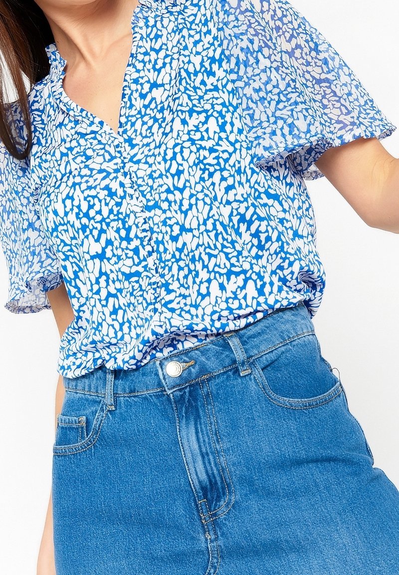 Blå blommig blus med korta volangärmar, med ett vitt abstrakt mönster. Matchad med högmidjade blå denimjeans.