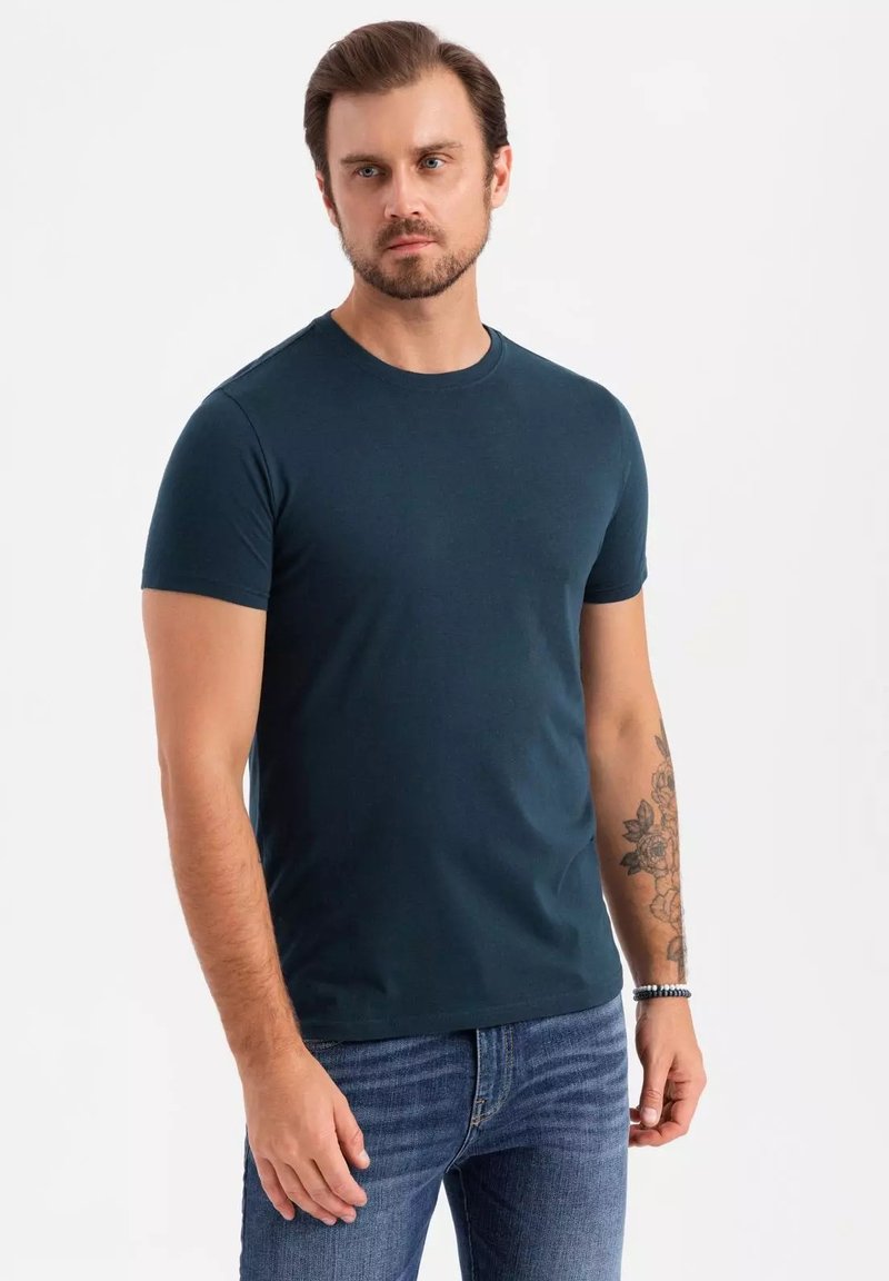 Mørkeblå kortærmet t-shirt med rund halsudskæring, lavet af blødt stof. Bæres med blå jeans. Har arm tatoveringer og et perlesmykke om håndleddet.