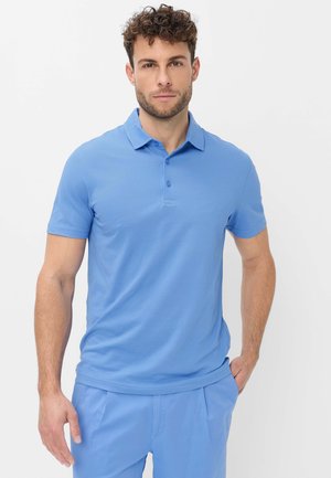 Man met krullend haar, gekleed in een lichtblauw poloshirt met korte mouwen en bijpassende broek, staand voor een effen witte achtergrond.