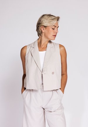 Blonde woman with short hair looks sideways, wearing a sleeveless beige vest over a white top and matching beige high-waist pants with hands in pockets.