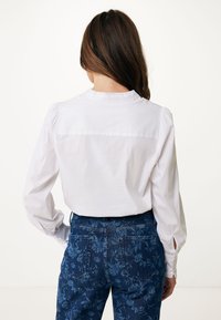 Camicia bianca a maniche lunghe con collo arricciato e bottoni. Abbinata a jeans blu scuro con motivi floreali e aperture laterali ai polsini.