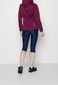 Bordeauxroter Kapuzenjacke, marineblaue knielange Leggings und marineblaue Sportschuhe. Die Jacke hat eine strukturierte Oberfläche und seitliche Reißverschlusstaschen.