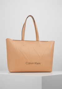 Tento lososově růžový kožený tote bag s embosovaným nápisem "NY" a "Calvin Klein" má dvě dlouhé úchyty a hladký povrch s minimalistickým designem.