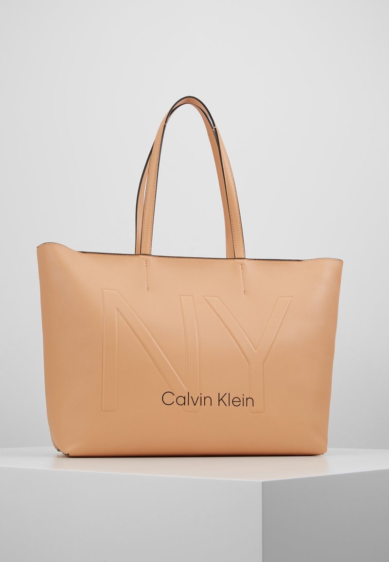 Tento lososově růžový kožený tote bag s embosovaným nápisem "NY" a "Calvin Klein" má dvě dlouhé úchyty a hladký povrch s minimalistickým designem.