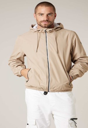 Man met kort haar, gekleed in een beige capuchonjas en witte cargobroek, staand met handen in de jaszakken.