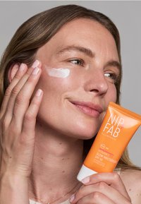 La crème hydratante NIP+FAB Vitamin C Fix Glow dans un tube orange. Un modèle applique la crème blanche sur sa joue, mettant en valeur une texture de peau lisse et un éclat naturel.