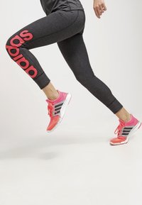 En person som har på sig mörkgrå Adidas leggings och rosa träningsskor i ett hopp mot en enfärgad ljus bakgrund.