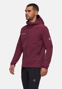 Homme portant une veste à capuche bordeaux avec fermeture éclair et un pantalon noir, se tenant debout et faisant face à gauche sur un fond blanc.