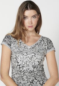 Tricou cu decolteu în V, de culoare gri, cu un imprimeu floral alb-negru. Mâneci scurte, fabricat dintr-un material moale, cu un design floral texturat în centru.