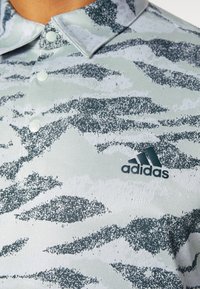 Polo shirt s límečkem v světle šedé barvě s zeleným digitálním maskáčovým vzorem; má tři knoflíky a černé logo Adidas na hrudi.