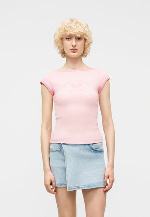 Blondynka o kręconych włosach, ubrana w dopasowany różowy top z krótkim rękawem i jasnoniebieską asymetryczną mini spódnicę dżinsową, stojąca przodem.