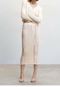 Frau in einem cremefarbenen, gerippten langen Rock und passendem Pullover mit dezenten Abnutzungsdetails, steht mit verschränkten Armen in einer neutralen Umgebung.