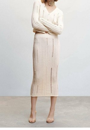 Kobieta w kremowej, prążkowanej spódnicy do kostek i dopasowanym swetrze z zniszczonymi detalami, stojąca z założonymi rękami w neutralnym otoczeniu.