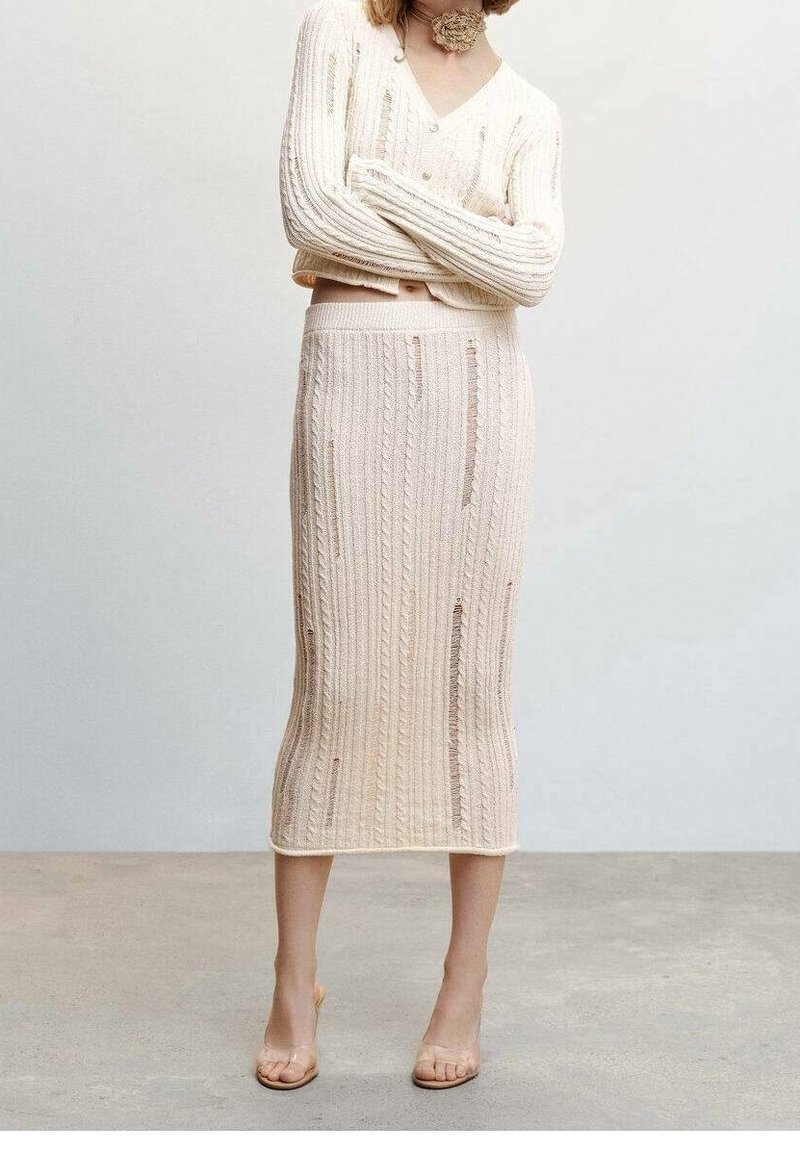 Frau in einem cremefarbenen, gerippten langen Rock und passendem Pullover mit dezenten Abnutzungsdetails, steht mit verschränkten Armen in einer neutralen Umgebung.