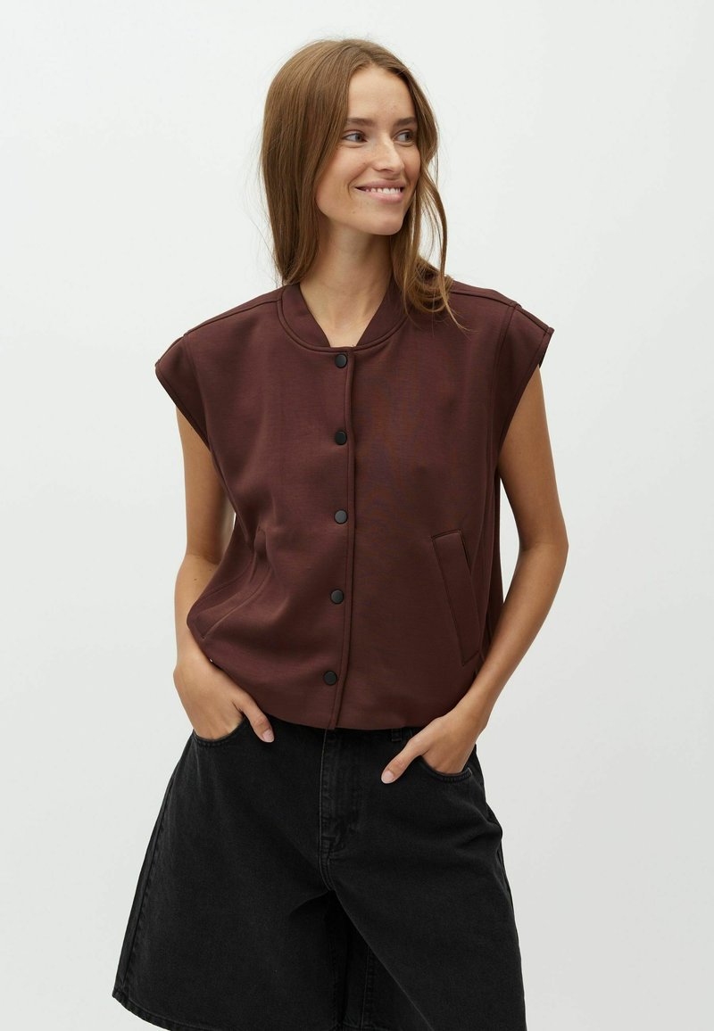 Jeune femme souriante de côté portant un gilet marron sans manches boutonné et un short noir ample sur un fond clair uni.