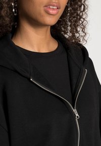Sudadera negra con cremallera hecha de una mezcla de algodón, que presenta una capucha, un cierre con cremallera y un acabado texturizado. Se puede ver una camiseta negra de manga corta debajo.