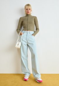 Jeans de cintura alta de um azul claro, combinados com um suéter cropped de zip-up ajustado na cor verde-oliva. Bolsa branca e ténis vibrantes com detalhes em rosa.