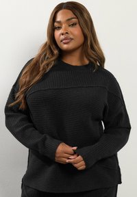 Svart ribbad stickad tröja med långa ärmar, avslappnad passform och rund halsringning. Har sidslitsar och ett strukturerat mönster genom hela plagget.