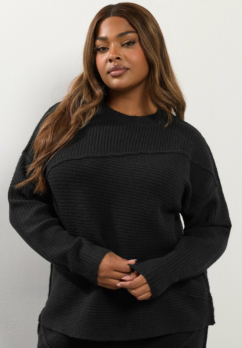 Svart ribbad stickad tröja med långa ärmar, avslappnad passform och rund halsringning. Har sidslitsar och ett strukturerat mönster genom hela plagget.
