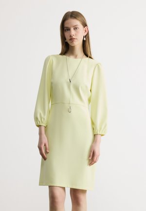 Mujer joven con vestido amarillo pálido hasta la rodilla, mangas largas abullonadas y un collar largo de plata, posando sobre un fondo blanco.