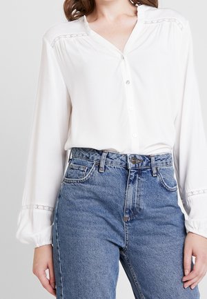 Kobieta ubrana w białą, długą bluzkę z koronkowymi detalami i wysokim stanem niebieskich dżinsów, ręce spoczywające wzdłuż ciała.