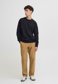 Melns džemperis ar apaļu kakla izgriezumu, bēšas krāsas bikses un baltas-melnas sporta kurpes. Modelis stāv pret gaiši pelēku fonu, viņam ir atslābināta poza.