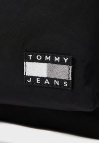 Juodas audinio lopas su tekstu "TOMMY" viršuje, po juo pilkai dryžuota ir balta juosta, o apačioje yra "JEANS", siuvinėta juodais siūlais.