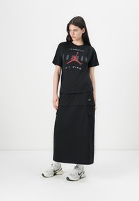 Černé tričko s červeným a šedým grafickým potiskem, doplněné černou maxi sukni. Obuv je bílá s zelenými akcenty. Jednoduchý design, atletický styl.