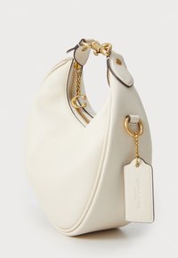 Borsa a tracolla in pelle crema a forma di mezza luna, con hardware dorato, chiusura con zip e un'etichetta rimovibile. Texture liscia con una leggera lucentezza.