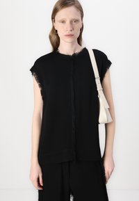 Svart ermeløs topp med frynsete kanter, laget av mykt stoff. Den er parret med en liten kremfarget skulderveske og har et enkelt, moderne design.