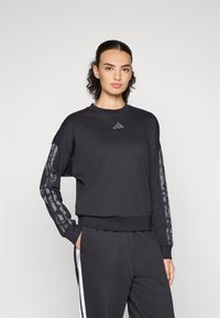Donna che indossa una felpa nera Adidas con strisce grigie a motivo sulle maniche e pantaloni neri con strisce bianche sui lati, in piedi contro uno sfondo bianco.