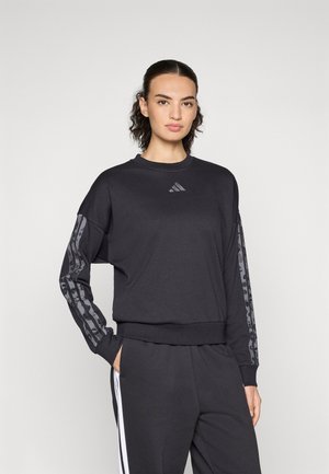 Femme portant un sweat à capuche noir Adidas avec des rayures grises à motifs sur les manches et un pantalon noir avec des rayures blanches sur les côtés, se tenant devant un fond blanc.