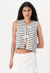 Gilet en tricot avec des rayures horizontales noir et blanc, col en V, devant ouvert et fermeture à nouer. Porté par-dessus une jupe blanche avec un ourlet à volants.